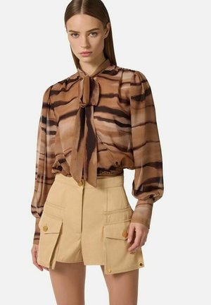 Donna che indossa una camicetta marrone a righe con collo a fiocco e maniche lunghe, abbinata a pantaloncini beige a vita alta con grandi tasche con bottoni.