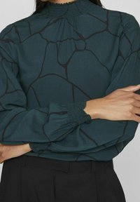 Blusa verde a maniche lunghe con scollo arricciato, caratterizzata da un motivo geometrico nero e polsini arricciati, indossata con pantaloni neri.