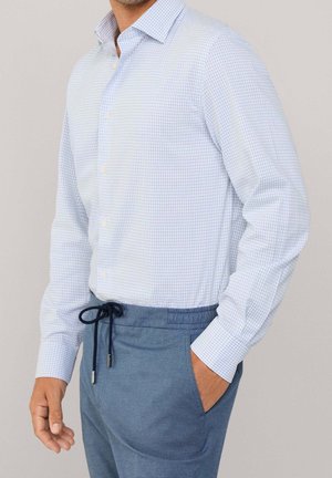 Homme portant une chemise à carreaux bleu clair rentrée dans un pantalon bleu à cordon, avec une main dans la poche et un fond neutre.
