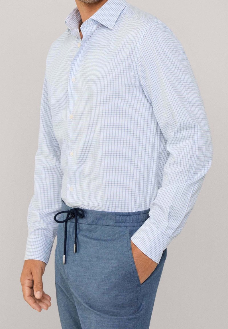 Homme portant une chemise à carreaux bleu clair rentrée dans un pantalon bleu à cordon, avec une main dans la poche et un fond neutre.