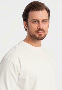 Uomo con capelli castani corti e barba curata, che indossa una maglietta bianca a girocollo semplice, guarda con calma la fotocamera su uno sfondo bianco.