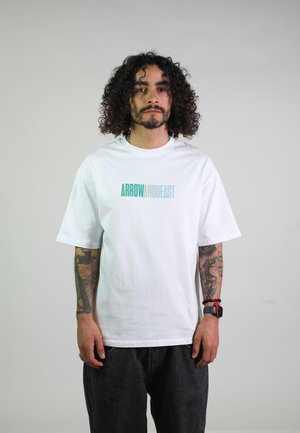 Mann mit lockigem Haar und Tattoos trägt ein weißes T-Shirt mit dem Schriftzug „ARROW AND BEAST“, schwarze Hose und eine Smartwatch mit rotem Armband.