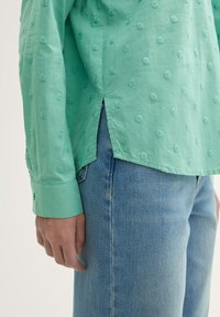 Camisa verde claro con patrón de puntos texturizados, aberturas laterales y puños abotonados, combinada con jeans de mezclilla azules. Ajuste casual y relajado.