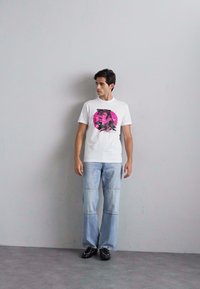 Fehér, rövid ujjú póló pink kör grafikával; világoskék, bőszárú farmernadrággal és fekete cipővel párosítva egy szürke háttér előtt.