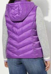Fialová péřová vesta s kapucí, s chevronovým prošíváním a lesklým povrchem. Nositelná přes šedý svetr s fialovými detaily.