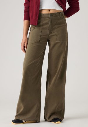 Femme portant un pantalon large vert olive avec de grandes poches avant, associé à un top blanc court et un cardigan bordeaux.