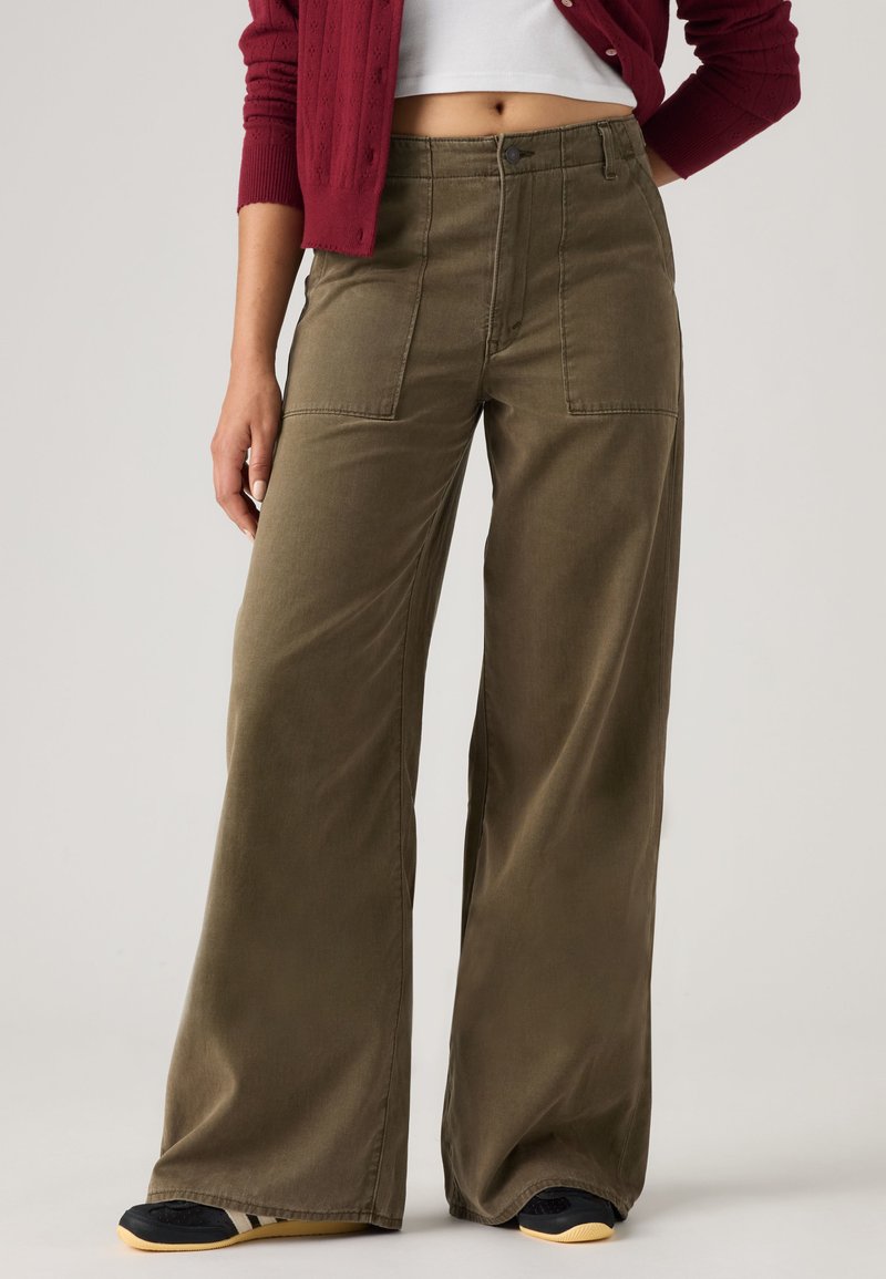 Femme portant un pantalon large vert olive avec de grandes poches avant, associé à un top blanc court et un cardigan bordeaux.
