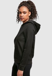 Black zip-up hoodie with a front pocket, curved hem, and a drawstring hood. Smooth texture with minimal detailing and a fitted design.
