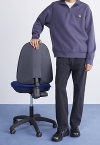 Henkilö, joka on pukeutunut purppuraan vetoketjulliseen huppariin ja mustiin housuihin, seisoo sinisellä lattialla, nojaten kättään mustan työtuolin selkänojaan, jossa on sininen istuin.