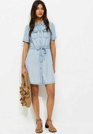 Vrouw in een lichtblauwe denimjurk met knopen en een taillekoord, met een strozak, en tankleurige sandalen met blokhak, staand tegen een witte achtergrond.