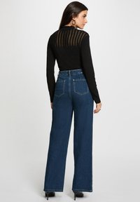 Femme portant un haut noir transparent à manches longues et un jean bleu taille haute à jambes larges avec des chaussures à talons noirs, se tenant devant un fond uni.