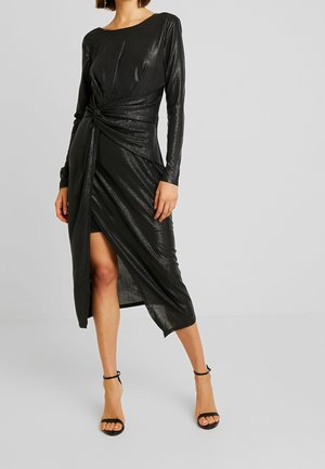 Kobieta ubrana w czarną sukienkę z długim rękawem, z detalem skręconego węzła i rozcięciem z przodu, w zestawie z czarnymi szpilkami na kostkę z paskiem.