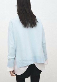 Svetlo modra majica z rebrastim robom in manšetami, ki ima sproščen kroj. Nosena prek bele srajce, z ukrivljenim zadnjim robom.