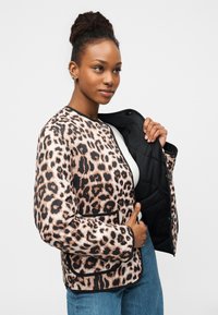 Kvinde med dreadlocks iført en jakke med leopardprint over en hvid top og blå jeans, holder jakken åben for at vise dens sorte foer.