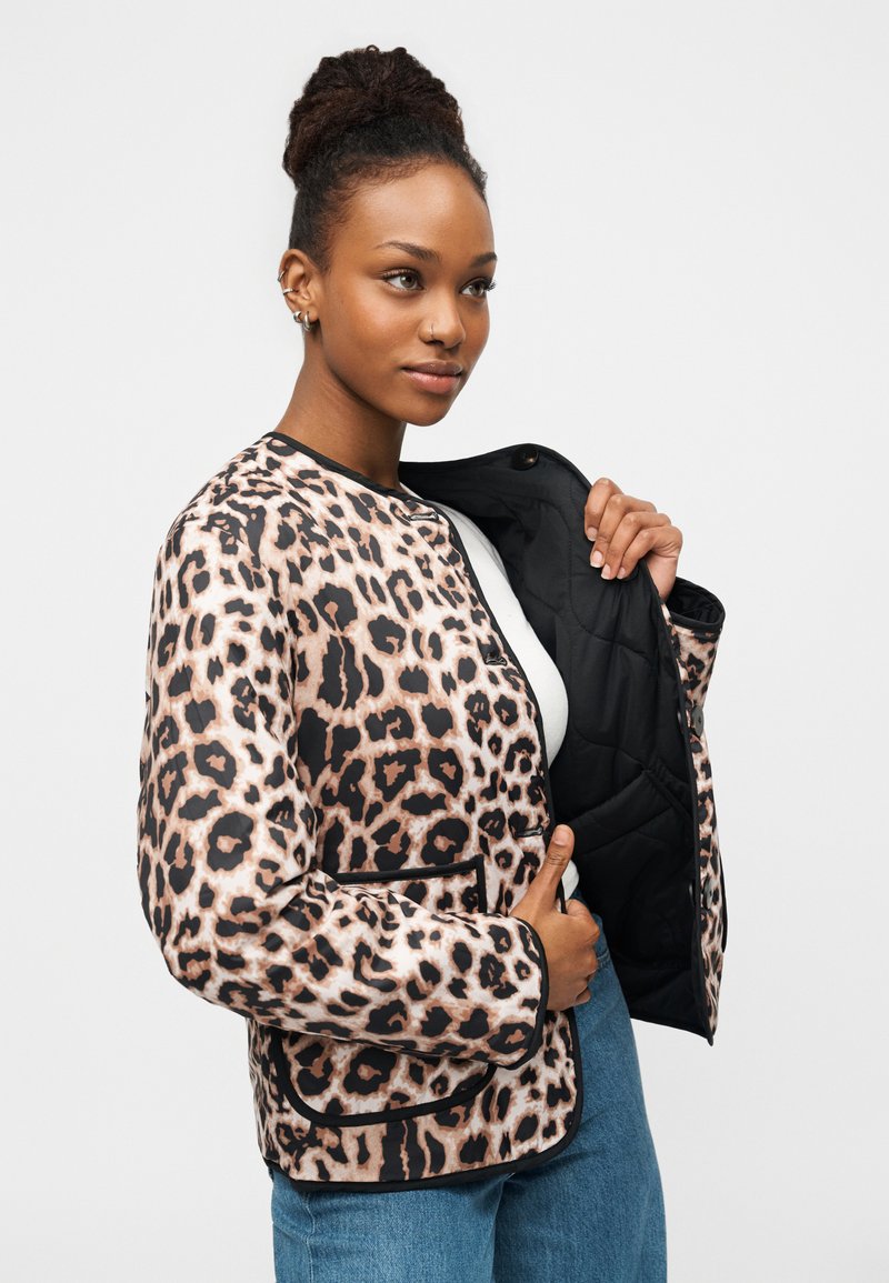 Kvinde med dreadlocks iført en jakke med leopardprint over en hvid top og blå jeans, holder jakken åben for at vise dens sorte foer.