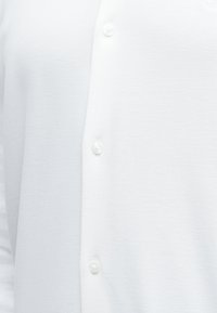 Κοντινό πλάνο λευκού πουκάμισου με υφή και τρία ορατά λευκά κουμπιά σε κάθετη γραμμή.