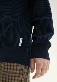 Granatowy sweatshirt o fakturze z ribbowym dekoltem i mankietami, z białą metką na boku. W komplecie z brązowymi spodniami w kratę.