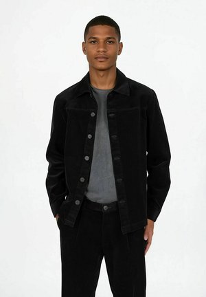 Ung mann iført svart jakke med knapper og bukser over en mørkegrå skjorte, stående med hendene i lommene mot en hvit bakgrunn.