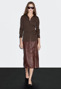 Braunes Button-Down-Hemd kombiniert mit einem braunen Leder-Midi-Rock. Das Model trägt übergroße Sonnenbrillen und schwarze spitz zulaufende Ballerinas.