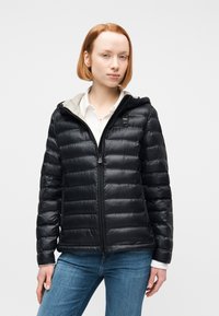 Νεαρή γυναίκα με κοντά κόκκινα μαλλιά, φοράει μαύρο καπιτονέ μπουφάν πάνω από λευκό πουκάμισο και μπλε τζιν, στέκεται μπροστά σε λευκό φόντο.