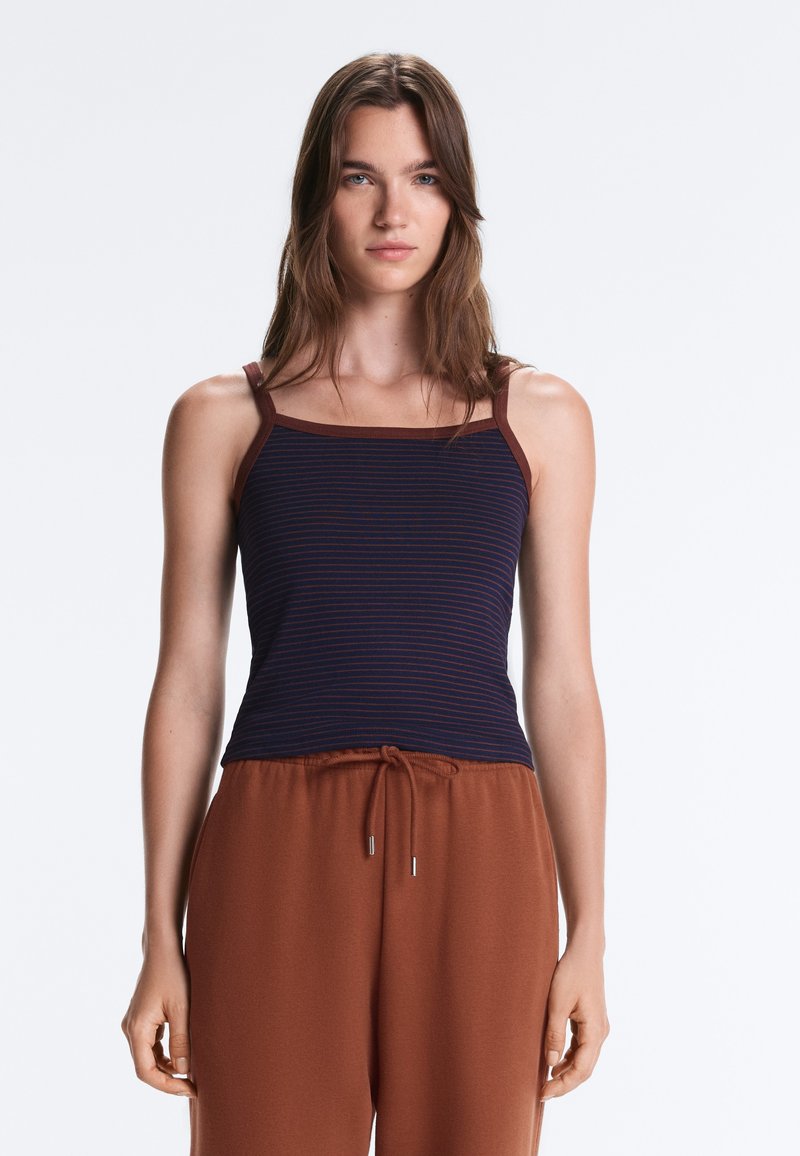 Jeune femme aux longs cheveux bruns portant un débardeur rayé bleu marine et un pantalon marron à cordon, debout devant un fond clair.