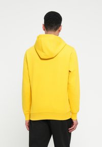 Muž s krátkymi kučeravými vlasmi, oblečený v jasne žltej mikine a čiernych nohaviciach, otočený chrbtom na jednoduchom bielom pozadí.