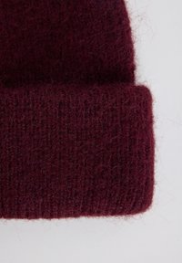 Bordeaux-neulottu pipo, jossa on pehmeä, teksturoitu pinta ja taitettu reunus. Materiaali näyttää pörröiseltä hienoilla ribbitiedoilla.