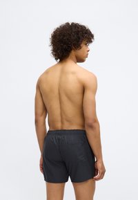 Melni peldēšanas šorti ar elastīgu jostasvietu un vienu aizmugures kabatu, kas izstrādāti no viegla, gluda auduma. Modelis redzams no aizmugures.