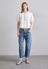Cămașă albă scurtă cu mâneci bufante, blugi deschiși la culoare cu talie înaltă și sandale deschise la culoare cu design cu bareta în T. Texturi netede pe toată suprafața.