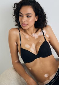 Black lace bra with a scalloped edge, adjustable straps, and prominent branding on the straps. Smooth surface and triangular cups.