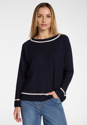 Frau mit glattem blondem Haar, die einen marineblauen Pullover mit weißem Zierstreifen an Kragen, Bündchen und Saum trägt, kombiniert mit blauer Jeans, stehend mit einer Hand in der Tasche.