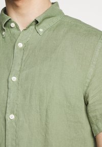 Vaaleanvihreä, lyhythihainen napitettava paita, joka on valmistettu kuvioidusta kankaasta. Sisältää kauluksen ja muoviset napit etuosassa.