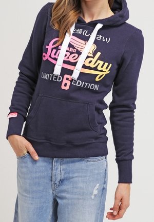 Kobieta w granatowej bluzie z kapturem z różowo-żółtym napisem "Superdry Limited Edition 6", ubrana w jasnoniebieskie podarte dżinsy, stojąca z ręką w kieszeni.