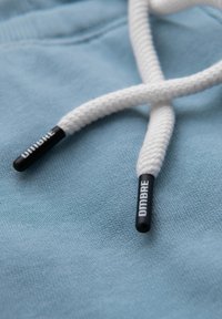 Tejido azul claro con una superficie texturizada, que presenta cordones de zapato trenzados en blanco con aglets negros marcados "DIMBRE". Diseño minimalista.