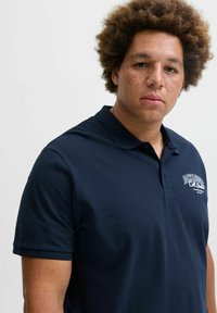 Dunkelblaues Polohemd aus strukturiertem Stoff, mit kurzen Ärmeln und Kragen. Mit weißen Logodetails auf der linken Seite.