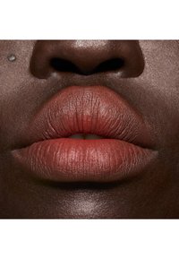 Gros plan sur des lèvres pleines, avec un rouge à lèvres mat de couleur brun rougeâtre. La peau a une texture lisse et foncée, et un petit grain de beauté se situe au-dessus de la lèvre.