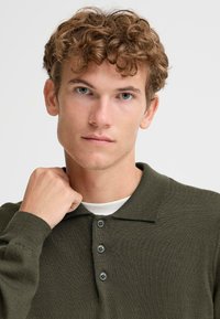Polo shirt in verde oliva lavorato a maglia con colletto strutturato, dotato di tre bottoni scuri. Indossato sopra una t-shirt bianca, che mette in evidenza un tessuto testurizzato.