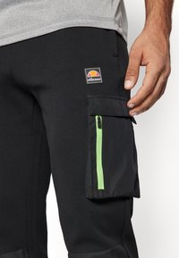Zwarte cargobroek met een groen ritsvak, een ellesse-logo en een slanke pasvorm. Het materiaal lijkt stevig met een gladde textuur.
