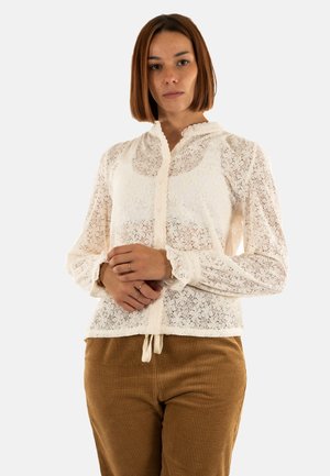 Blouse en dentelle blanche à manches longues, fermeture par boutons, motif texturé et détail de col froncé, associée à un pantalon en velours côtelé marron.