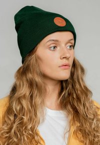 Zielona dziana czapka beanie z brązową skórzaną naszywką z motywem górskim, noszona przez osobę z długimi, falującymi włosami na neutralnym tle.