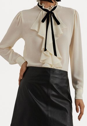 Femme portant un chemisier crème à volants avec un ruban noir et une jupe en cuir noire en ligne A, main posée sur la hanche.