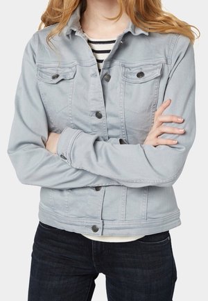 Personne aux bras croisés portant une veste en jean gris clair par-dessus une chemise rayée noire et blanche et un jean foncé.
