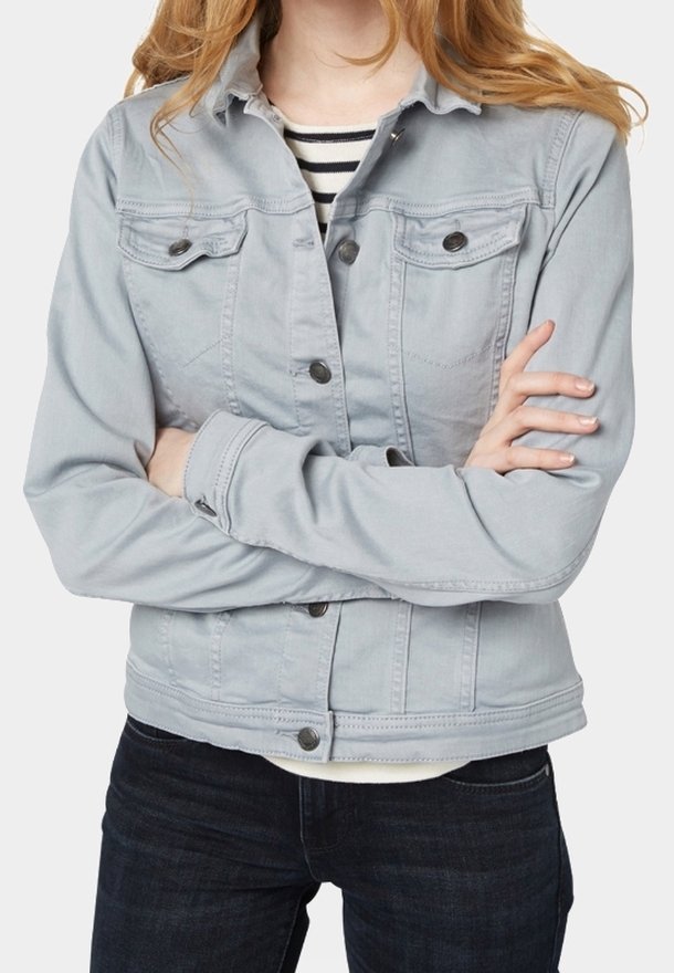 Personne aux bras croisés portant une veste en jean gris clair par-dessus une chemise rayée noire et blanche et un jean foncé.
