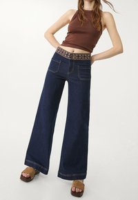 Nainen, jolla on tummat leveät farkut, joissa on kirjailtu vyötärö, ja ruskea ilmanhihoin crop-top, poseeraa kädet lanteilla.