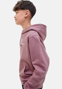 Une jeune personne avec des cheveux courts et stylés est montrée de profil, portant un hoodie violet avec un logo, les mains dans la poche. Fond simple.