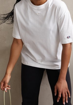 Osoba ubrana w białą koszulkę Champion z krótkim rękawem, z logo na klatce piersiowej i rękawie, trzymająca w lewej ręce jasnziezielony przedmiot.