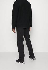 Schwarzer Fleece-Pullover, lockere Passform, lange Ärmel. Kombiniert mit dunklen Jeans und Sneakers mit einem schwarz-weißen gepunkteten Sohlenmuster.