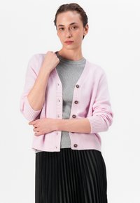 Cárdigan rosa claro con puños y dobladillo acanalados. Abrochado con botones oscuros, superpuesto a una camisa gris y una falda negra plisada.
