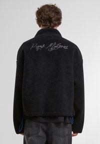 Veste en polaire noire avec un col haut, présentant un texte brodé au dos. Comprend des accents bleus au niveau de l'ourlet.