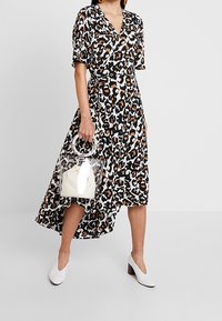 Leopardmönstrad omslagsklänning med korta ärmar, hög-låg fåll och en färgpalett i svart, beige och brunt. Matchad med vita skor och en transparent väska.